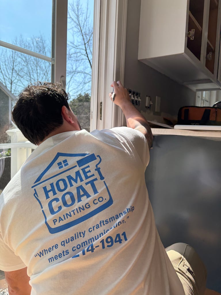 Worker in Home Coat Painting Co. shirt standing at kitchen cabinets by a window
